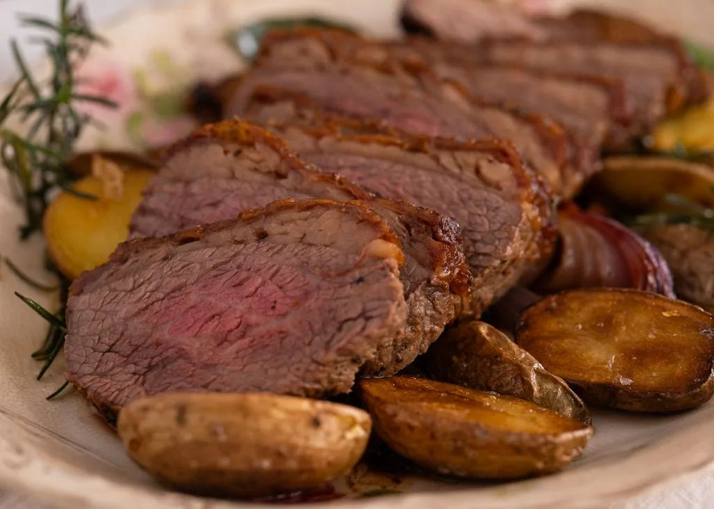 Carne assada no forno fatiada, rosada no centro, servida com batatas assadas, cebola roxa e alecrim